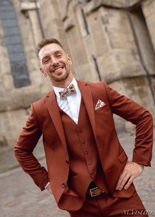 Le marié se tient fièrement devant une église ancienne de Romans, reconnaissable à sa façade en pierre beige et ses grandes fenêtres en ogive. Il porte un costume terracotta, lumineux et moderne, assorti à un gilet de la même teinte. Sa chemise blanche contraste élégamment avec l’ensemble, tandis que son nœud papillon et sa pochette à motifs floraux dans les tons rose, vert et beige apportent une touche joyeuse et raffinée. Son sourire et son attitude détendue renforcent l’ambiance chaleureuse de cette scène de mariage en ville.