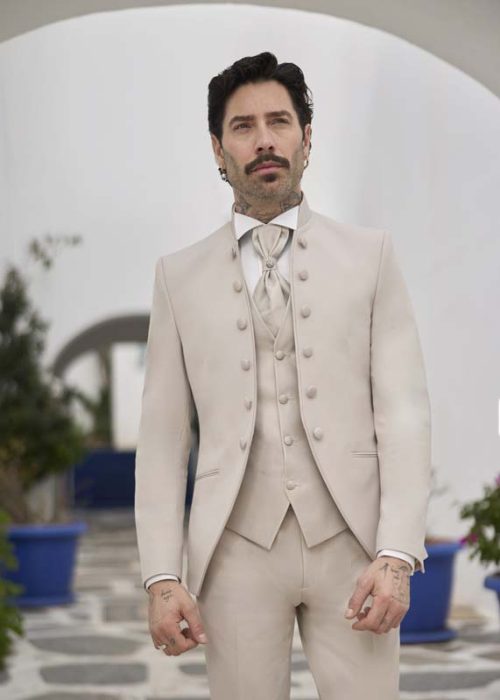 CADIX BEIGE_1520_optim-scaled L’homme porte un costume de cérémonie beige clair, très élégant et structuré. La veste à col montant se distingue par une rangée de boutons ton sur ton, renforçant un style à la fois classique et sophistiqué. Le gilet assorti et la cravate lavallière beige perlé, ornée d’une broche argentée, apportent une touche encore plus raffinée. La chemise blanche crée un contraste net et lumineux. L’ensemble dégage une allure noble, épurée et très cérémoniale.