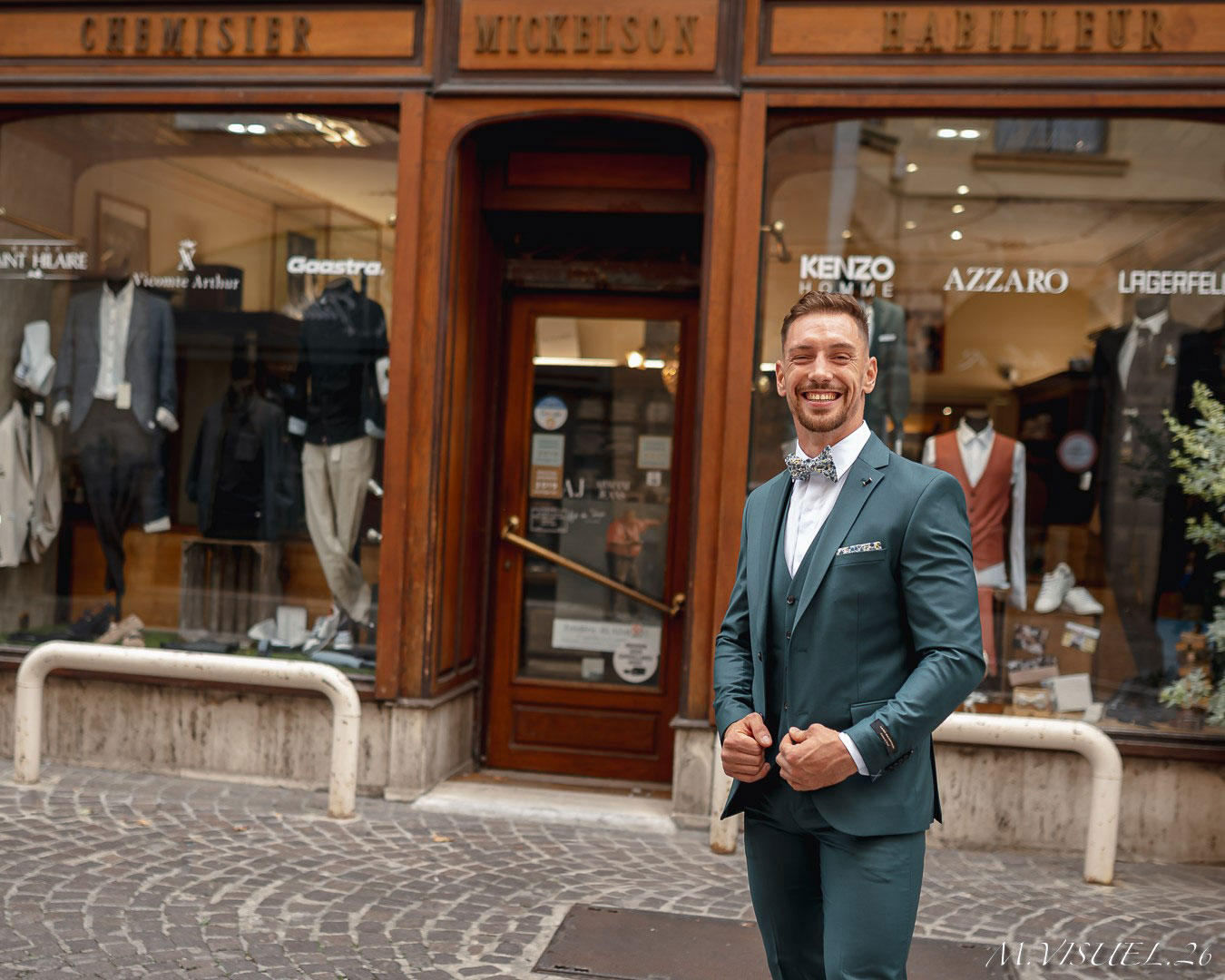 L’homme pose avec assurance devant Mickelson, le magasin spécialisé en costumes pour hommes et tenues de mariés situé au cœur de Romans. Il porte un costume vert pétrole parfaitement ajusté, assorti d’un gilet et d’un nœud papillon à motifs, créant un look moderne et élégant. La boutique, avec sa façade en bois et ses vitrines bien fournies, expose des marques prestigieuses comme Kenzo, Azzaro, Lagerfeld ou encore Victoire Arthur, soulignant son expertise dans l’habillement masculin haut de gamme. Le sol pavé et l’ambiance citadine donnent un charme authentique à la scène, mettant en valeur l’élégance du costume et le savoir-faire de la maison Mickelson.
