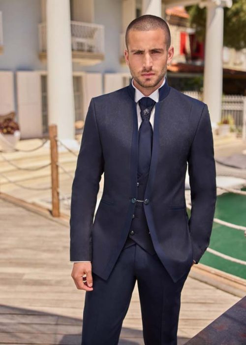 sao-paulo-marine-scaled L’homme porte un costume trois-pièces bleu marine au style très élégant et structuré, avec une veste à col montant qui modernise l’ensemble. Le gilet assorti est fermé par une chaîne décorative discrète, renforçant le côté raffiné. La cravate bleu marine, ornée d’un nœud décoratif sombre, ajoute une touche formelle et distinguée. La chemise blanche crée un contraste net et propre. L’ensemble offre une silhouette chic, équilibrée et résolument habillée.