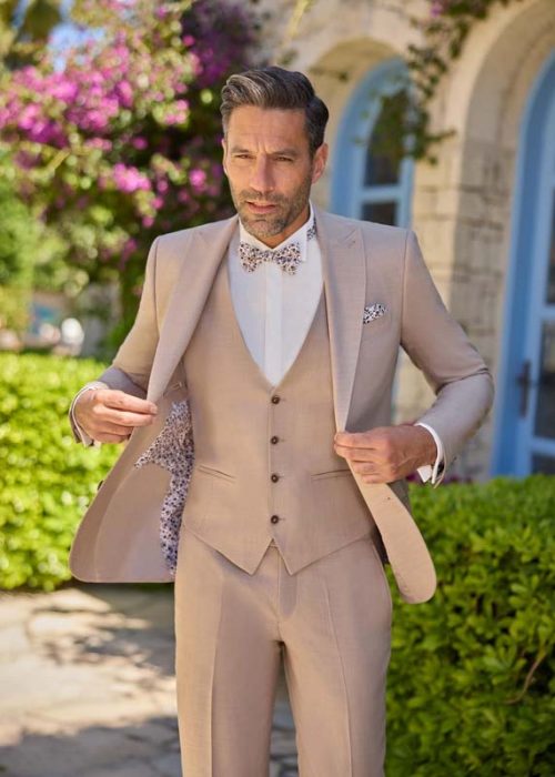 sienne-sable-scaled Un homme porte un costume trois-pièces élégant de couleur beige clair sienne tirant vers le sable, parfaitement ajusté. Il ouvre sa veste pour laisser apparaître une doublure à motif floral assortie au nœud papillon et à la pochette. La chemise blanche apporte un contraste net et raffiné à l’ensemble. Le style est chic, lumineux et idéal pour une ambiance estivale. L’environnement extérieur fleuri renforce l’atmosphère élégante et chaleureuse de la scène.