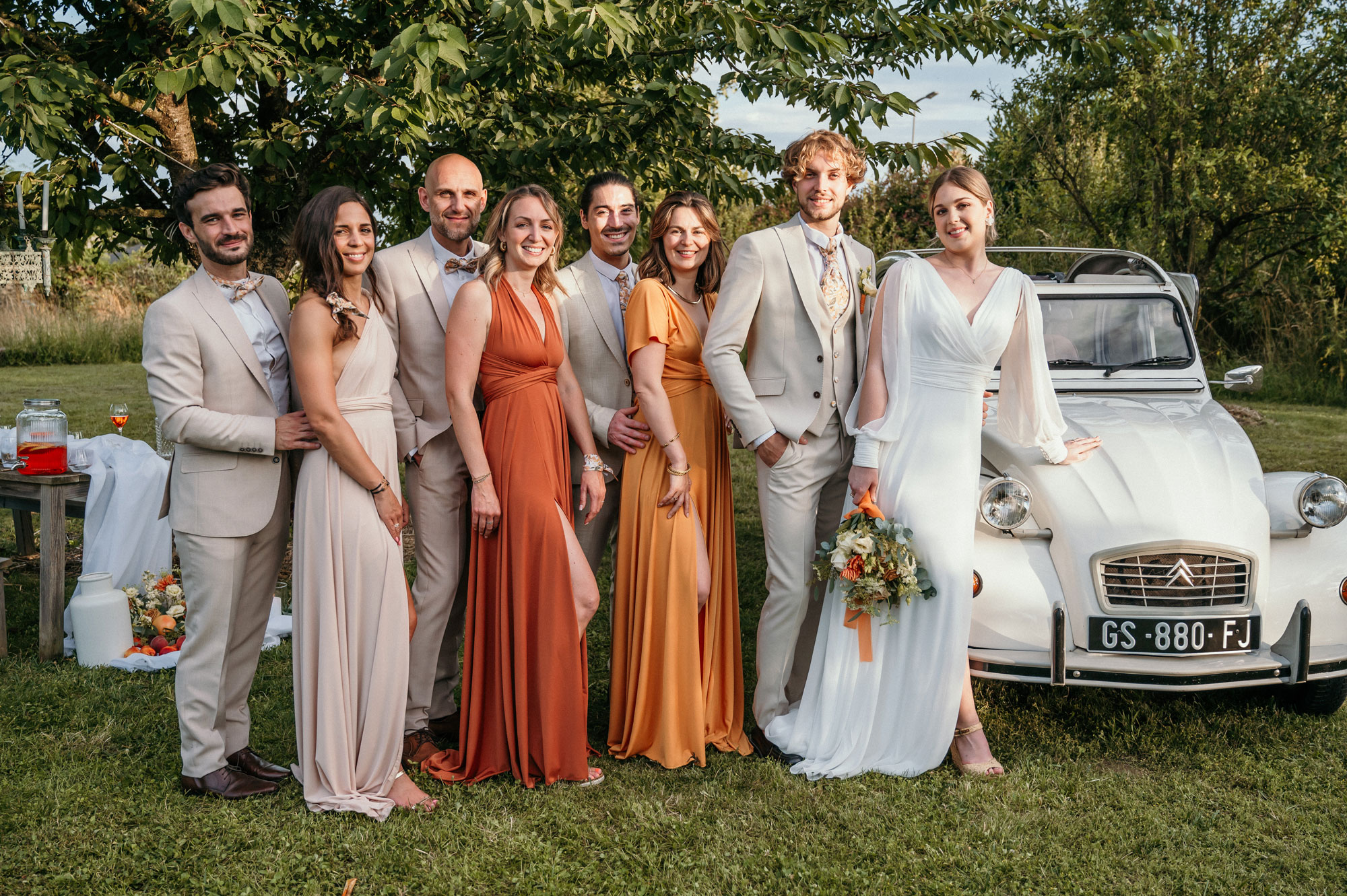 Mariage champêtre en extérieur, ambiance bohème chic avec une voiture rétro blanche. Palette très naturelle : beige/sable/lin, avec des touches chaudes terracotta, rouille et ocre. Les hommes portent des costumes beige clair, coupe ajustée, chemise claire et accessoires floraux. Le marié est en costume beige avec gilet et cravate fleurie assortie aux tons orangés. Les femmes ont des robes longues fluides terracotta/nude/ocre, la mariée en robe ivoire avec bouquet eucalyptus et orange. Costume Mickelson