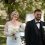 Mariés souriants en extérieur lors d’un mariage. La mariée porte une robe blanche avec bouquet clair, tandis que le marié porte un élégant smoking noir avec nœud papillon et fleur blanche à la boutonnière, dans un cadre verdoyant et lumineux. Costume Mickelson à Romans
