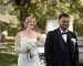 Mariés souriants en extérieur lors d’un mariage. La mariée porte une robe blanche avec bouquet clair, tandis que le marié porte un élégant smoking noir avec nœud papillon et fleur blanche à la boutonnière, dans un cadre verdoyant et lumineux. Costume Mickelson à Romans