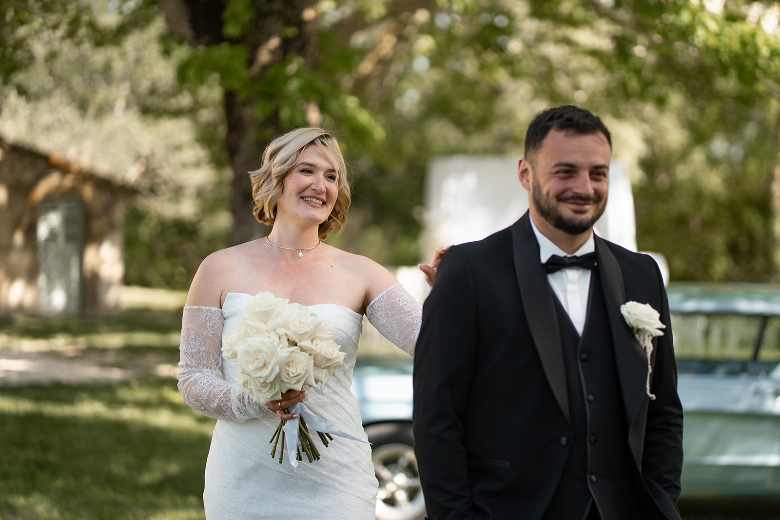 Mariés souriants en extérieur lors d’un mariage. La mariée porte une robe blanche avec bouquet clair, tandis que le marié porte un élégant smoking noir avec nœud papillon et fleur blanche à la boutonnière, dans un cadre verdoyant et lumineux. Costume Mickelson à Romans