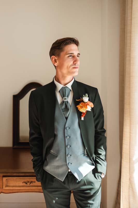 Dans la lumière naturelle d’une élégante chambre, ce costume trois-pièces vert profond se distingue par l’harmonie parfaite de ses détails. Le gilet gris perle, la lavallière assortie et la boutonnière aux nuances orangées apportent une touche de raffinement et de caractère à l’ensemble. La coupe ajustée souligne une silhouette élégante et contemporaine, idéale pour une cérémonie d’exception. Une tenue sophistiquée qui conjugue tradition, modernité et élégance intemporelle.