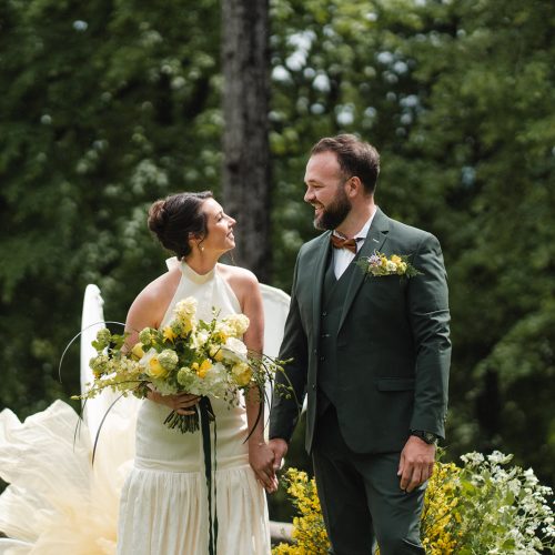 Main dans la main, les mariés avancent sous les regards de leurs invités dans un décor naturel et lumineux. Le marié porte un élégant costume trois-pièces vert foncé signé Mickelson, rehaussé d’un nœud papillon cuivré et d’une boutonnière florale aux tons jaunes et verts. La coupe ajustée met en valeur une silhouette moderne et raffinée, parfaitement en harmonie avec l’esprit champêtre de la cérémonie. Les sourires échangés et les couleurs coordonnées de leurs tenues créent une scène pleine d’émotion et d’élégance.