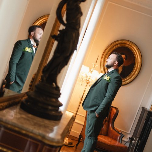 Dans l’élégance feutrée d’un intérieur de caractère, le marié prend la pose avec assurance dans son costume trois-pièces vert foncé signé Mickelson. Son nœud papillon cuivré, sa boutonnière florale et ses chaussures en cuir cognac apportent une touche de distinction parfaitement maîtrisée. Le jeu de miroir et la lumière chaleureuse mettent en valeur la qualité de la coupe et les détails du costume. Une silhouette raffinée et contemporaine qui s’intègre avec naturel dans ce décor intemporel.