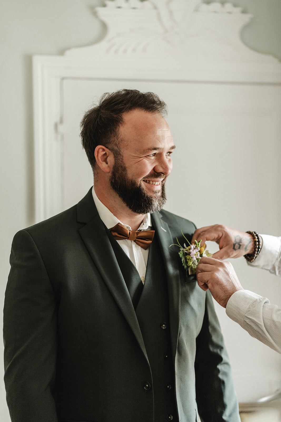 Dans les derniers préparatifs avant la cérémonie, le marié affiche un large sourire tandis que sa boutonnière est soigneusement ajustée. Son costume trois-pièces vert foncé signé Mickelson, associé à un nœud papillon cuivré, révèle toute son élégance et son originalité. Les détails floraux, harmonieusement accordés au thème du mariage, apportent une touche de fraîcheur et de raffinement. Une tenue de caractère qui conjugue parfaitement modernité, distinction et émotion.