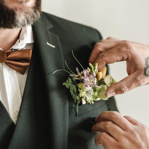 Gros plan sur les détails qui font toute la différence : le costume trois-pièces vert foncé signé Mickelson est sublimé par un élégant nœud papillon cuivré et une boutonnière florale délicatement travaillée. Les nuances de vert, de jaune et de violet rappellent l’univers végétal du mariage et apportent une touche de fraîcheur à l’ensemble. La qualité de la coupe et le soin apporté aux finitions soulignent l’élégance naturelle du marié. Une tenue raffinée, pensée jusque dans les moindres détails.