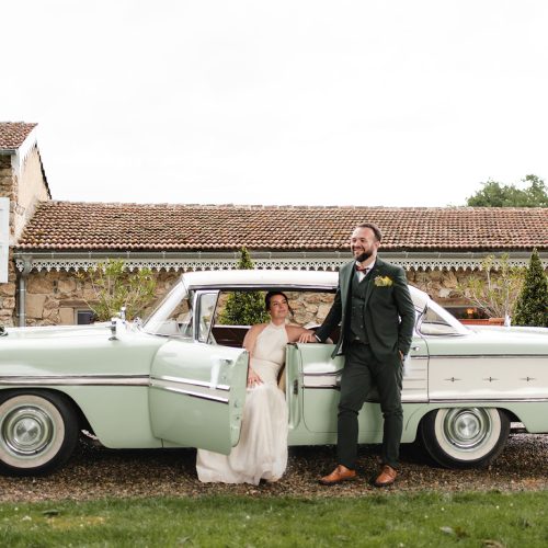 Devant une élégante voiture américaine de collection aux lignes emblématiques, le marié affiche un style affirmé dans son costume trois-pièces vert foncé acheté chez Mickelson. Son nœud papillon cuivré, sa boutonnière fleurie et ses chaussures en cuir cognac apportent une touche d'originalité et de raffinement à l'ensemble. Adossé à la portière ouverte, il pose avec assurance tandis que la mariée prend place à bord. Le contraste entre le costume contemporain et le charme vintage du véhicule crée une scène de mariage à la fois élégante et intemporelle.