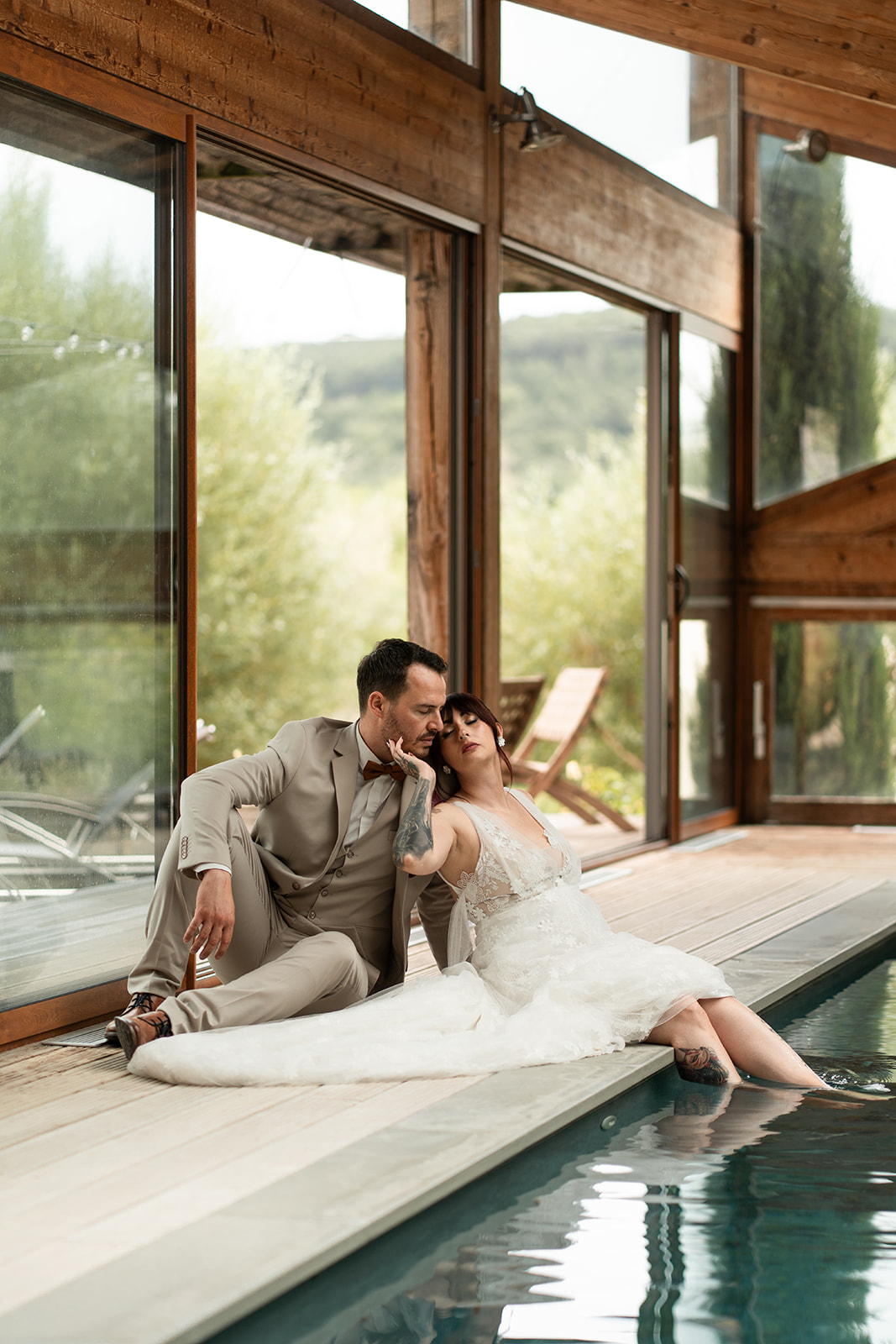 Assis au bord de la piscine, les mariés partagent un moment de calme et de complicité dans un cadre élégant baigné de lumière naturelle. Le costume beige du marié, associé à un nœud papillon cognac ton sur ton, apporte une allure à la fois chic et décontractée. Sa coupe ajustée et son gilet assorti soulignent une silhouette moderne, parfaitement adaptée à l’atmosphère raffinée du lieu. Une tenue élégante qui accompagne avec subtilité la douceur et l’intimité de cette scène.