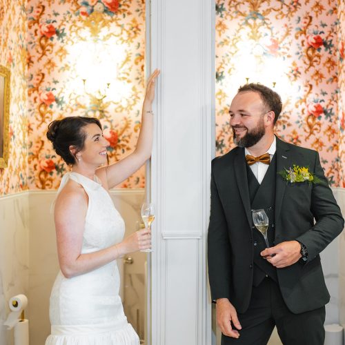 Le marié porte un élégant costume trois-pièces vert foncé, sublimé par un nœud papillon cuivré et une boutonnière florale aux tons verts et jaunes. Séparé de la mariée par une cloison centrale, il échange avec elle un regard complice tout en tenant une flûte de champagne. La coupe ajustée du costume met en valeur une silhouette raffinée et contemporaine. Ce costume, acheté chez Mickelson, apporte une touche de caractère et d’élégance à cette scène de mariage au décor romantique et chaleureux.
